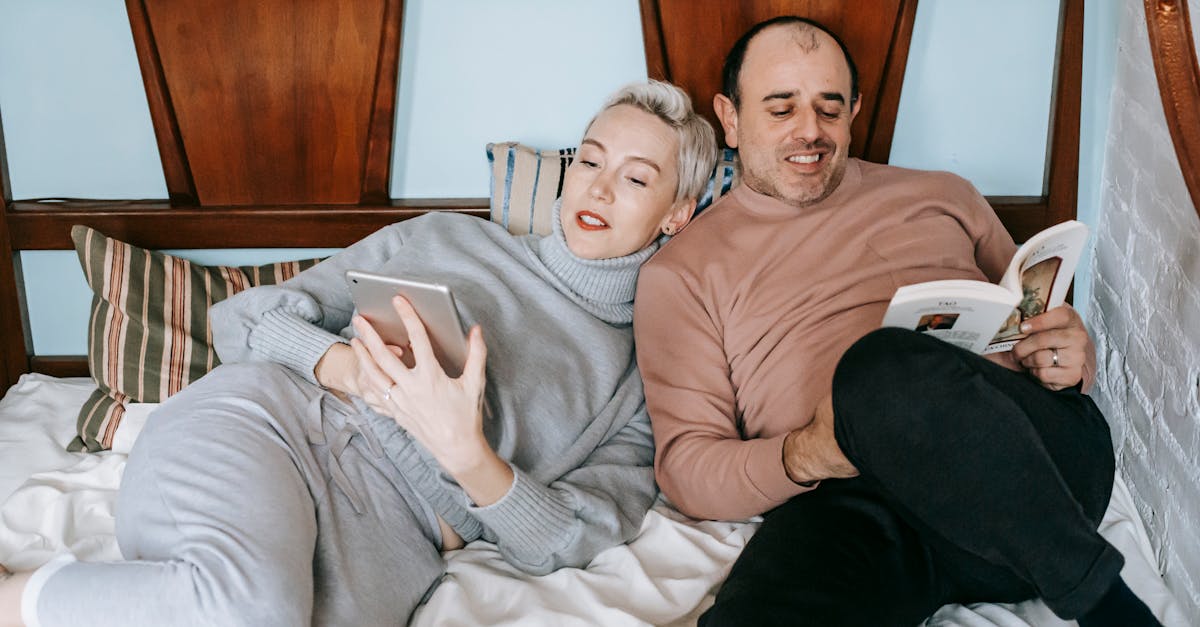 Happy middle aged multiethnic couple cuddling while lying on comfortable bed together with book and tablet during lazy weekend at home: marriage in the empty nest years